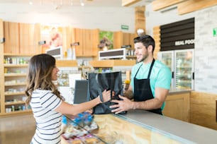 Cajero masculino seguro de sí mismo dando bolsa a la mujer en el mostrador de pago en la tienda de comestibles