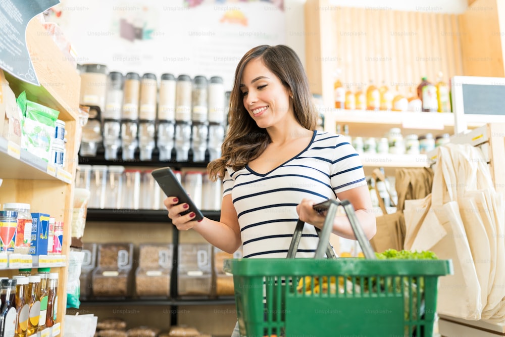 Mujer caucásica sonriente usando un teléfono inteligente mientras compra productos en la tienda de comestibles
