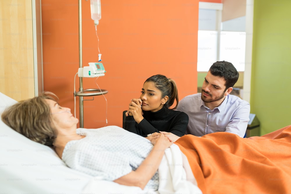 病院でベッドのそばに座りながら年配の母親を見ている悲しい夫婦