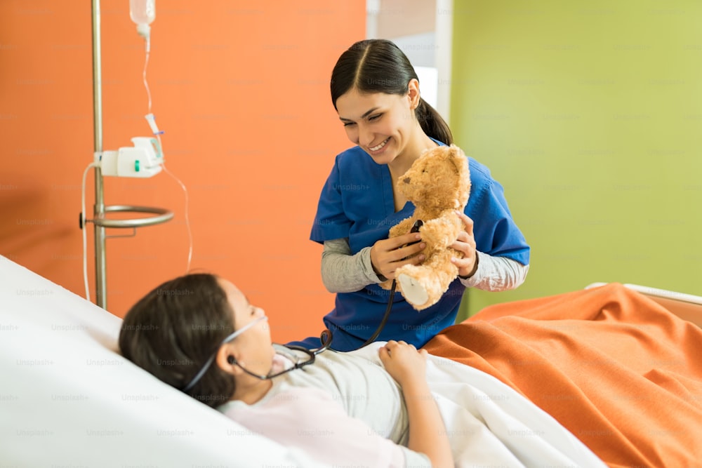 Young nurse and girl playing while holding teddy bear and stethoscope at hospital
