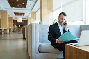 Handsome mid adult businessman busy on call while reading business plan from file in hotel lobby