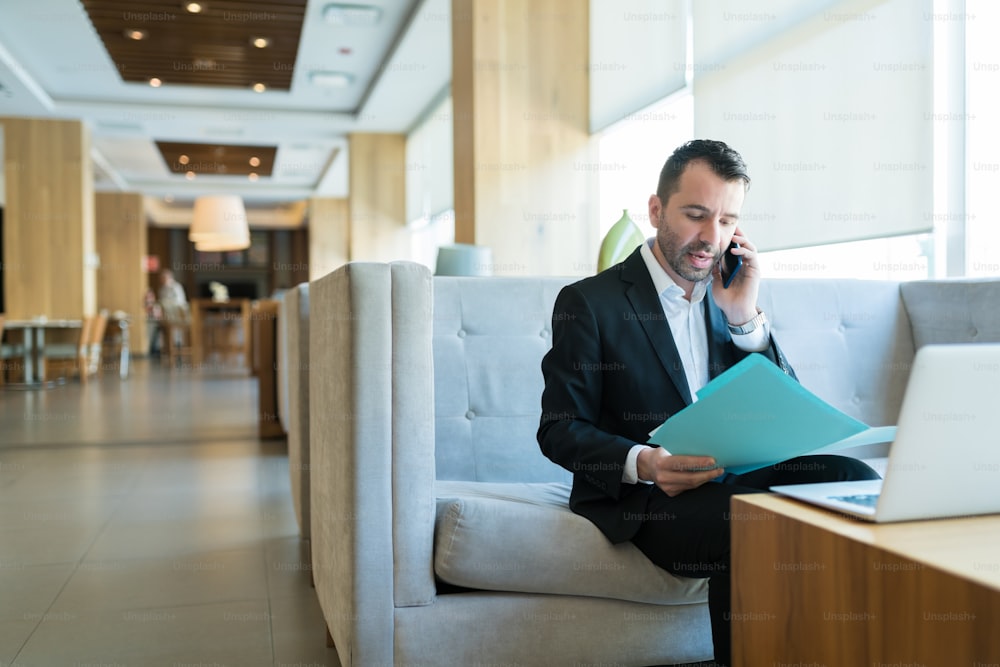 Hübscher Geschäftsmann mittlerer Erwachsener, der auf Abruf beschäftigt ist, während er den Geschäftsplan aus der Akte in der Hotellobby liest