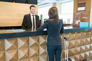 Guapo gerente adulto medio sonriendo mientras mira a la mujer de negocios de pie con equipaje en la recepción del hotel