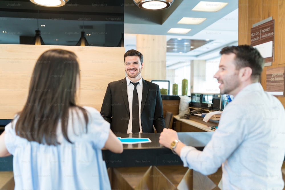 Handsome Hispanic manager welcoming newlywed couple on honeymoon trip while standing at front desk in hotel