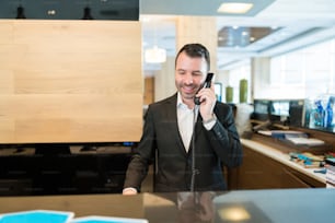 Happy male receptionist confirming booking while talking on phone at front desk in hotel