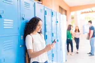 Smiling teenage student texting on mobile phone by lockers in high school