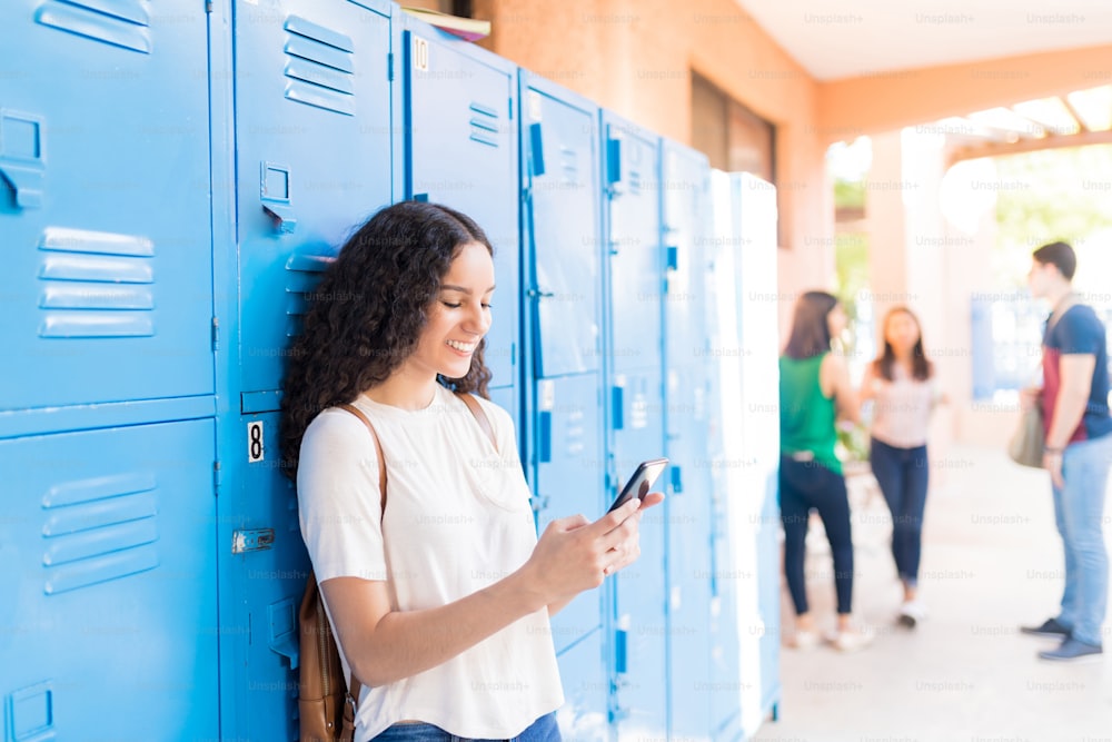 Smiling teenage student texting on mobile phone by lockers in high school