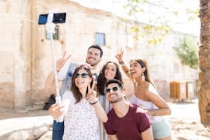 Happy male and female friends showing victory gesture while taking selfie on smart phone during summer weekend