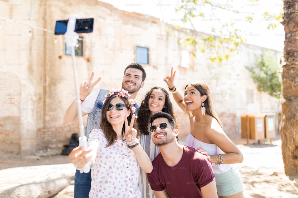 Des amis heureux hommes et femmes montrant un geste de victoire tout en prenant un selfie sur un téléphone intelligent pendant le week-end d’été