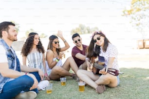 Amigos sonrientes escuchando a una mujer tocando la guitarra mientras disfruta de bebidas en un festival de música