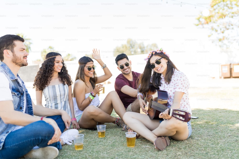 Smiling friends listening to woman playing guitar while enjoying drinks at music festival