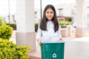 Retrato de una niña adorable sonriendo mientras lleva un bote de basura en el jardín