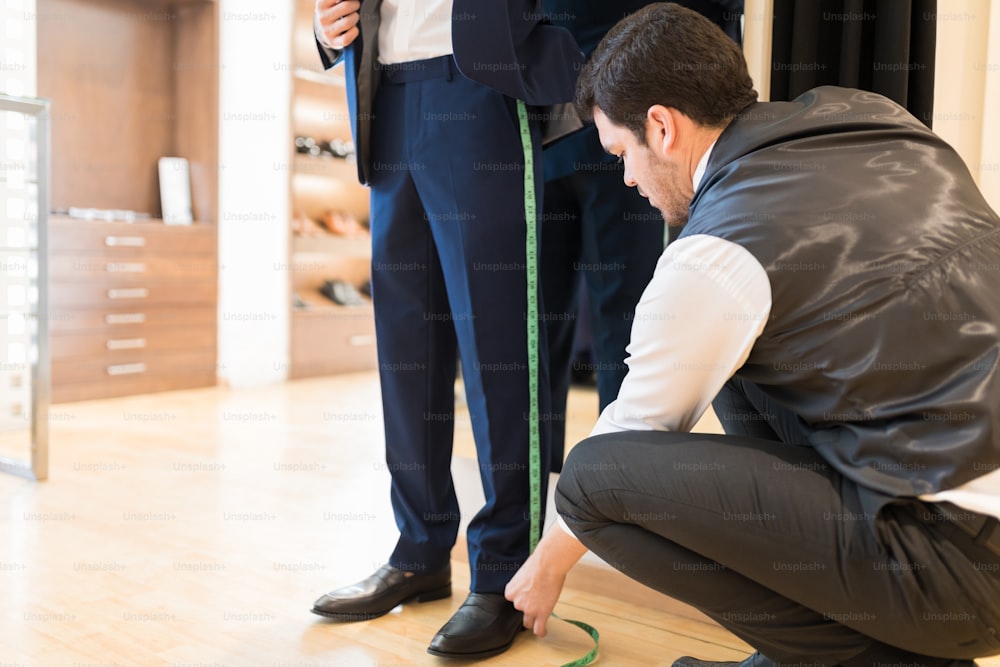Young Hispanic garment-maker measuring male customer's leg at tailor shop