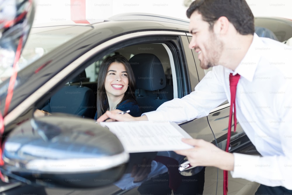 Salesman with financial document talking to client sitting in new car at showroom