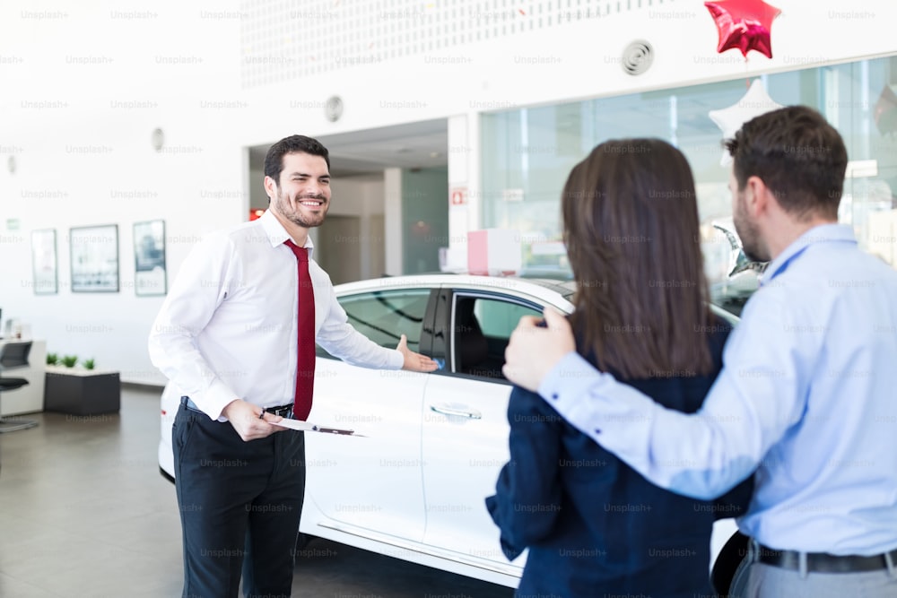 Good looking salesperson presenting brand new car to couple at showroom