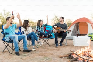 Fun loving camping friends singing together while sitting with man playing guitar