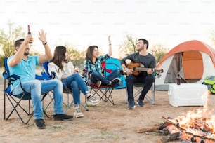 Amigos de camping amantes de la diversión cantando juntos mientras están sentados con un hombre tocando la guitarra