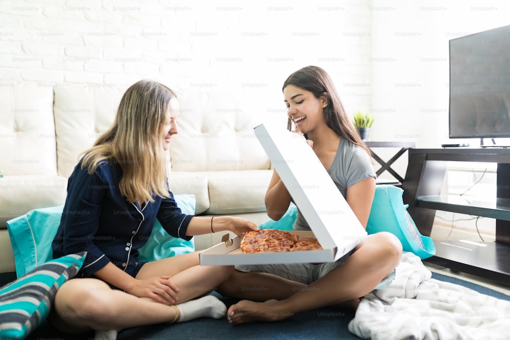거실에 앉아 있는 동안 친구를 위해 피자 상자를 여는 행복한 젊은 여자
