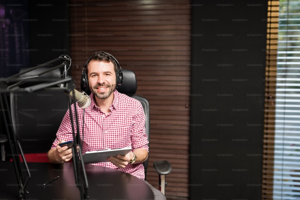 Retrato de un sonriente presentador de radio hispano con portapapeles presentando un programa en una estación de radio