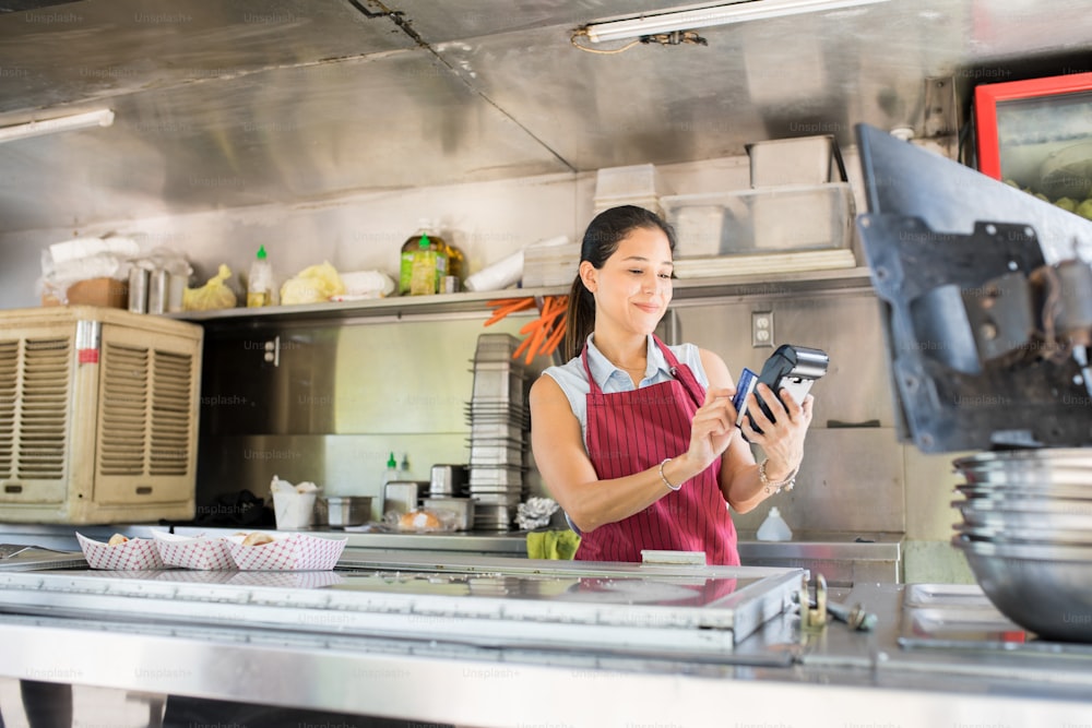 Jolie jeune femme travaillant dans un camion de nourriture et glissant une carte de crédit pour traiter un paiement
