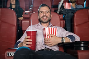 Joven feliz con barba abrazando una combinación de refrescos y palomitas de maíz mientras ve una película en el cine