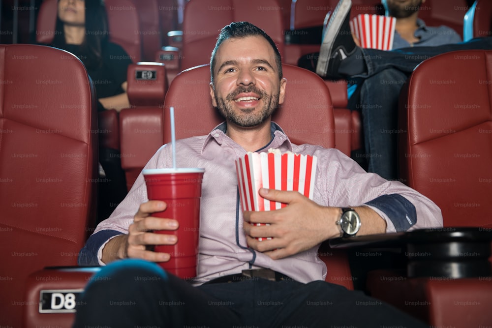 Jovem feliz com barba abraçando um combo de refrigerante e pipoca enquanto assiste a um filme no cinema