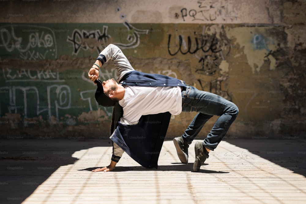 Jovem dançarino de hip hop masculino arqueando para trás e mostrando alguns de seus movimentos de dança em um cenário urbano com paredes de grafite