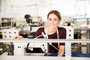 Ritratto di una splendida giovane sarta seduta davanti a una macchina da cucire in una fabbrica tessile e sorridente