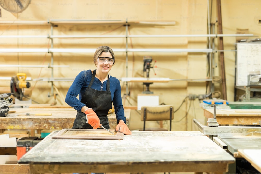 Portrait of a cute Hispanic female carpenter working on a cabinet door, using a wood gouge in her woodshop