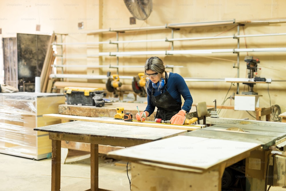 Wide view of a busy female carpenter measuring and marking some wood in her workshop
