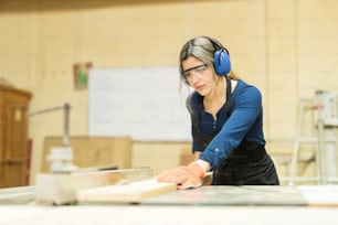 Good looking young female carpenter wearing earmuffs and using a table saw at work