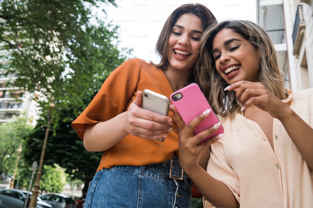 Two young friends enjoying together and using their mobile phones while standing outdoors on the street. Urban concept.