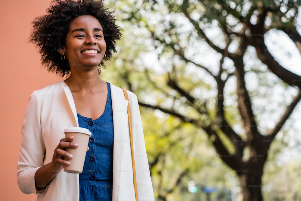 Ritratto di donna d'affari afro sorridente e tenendo in mano una tazza di caffè mentre si trova all'aperto sulla strada. Concetto commerciale e urbano.