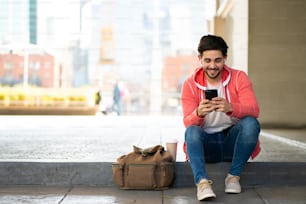 Retrato de un joven usando su teléfono móvil mientras está sentado al aire libre en la calle. Concepto urbano.