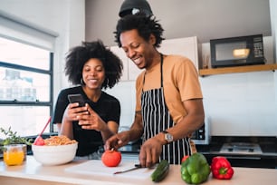 Ritratto di giovane coppia afro che cucina insieme e usa il telefono cellulare nella cucina di casa. Concetto di relazione, cucina e stile di vita.