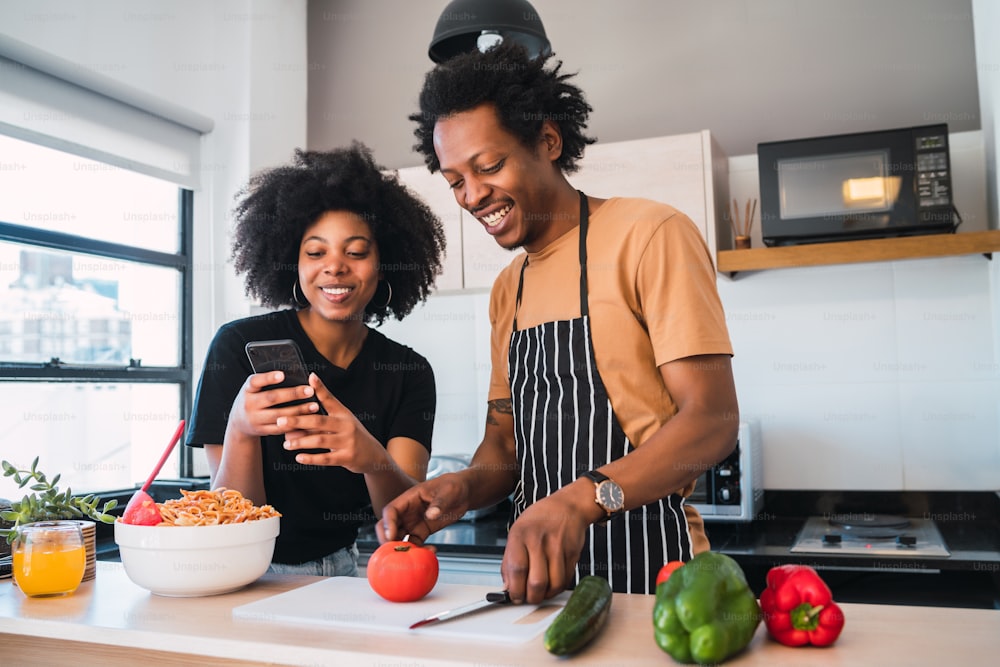 Retrato de jovem casal afro cozinhando juntos e usando o telefone celular na cozinha de casa. Relacionamento, cozinheiro e conceito de estilo de vida.