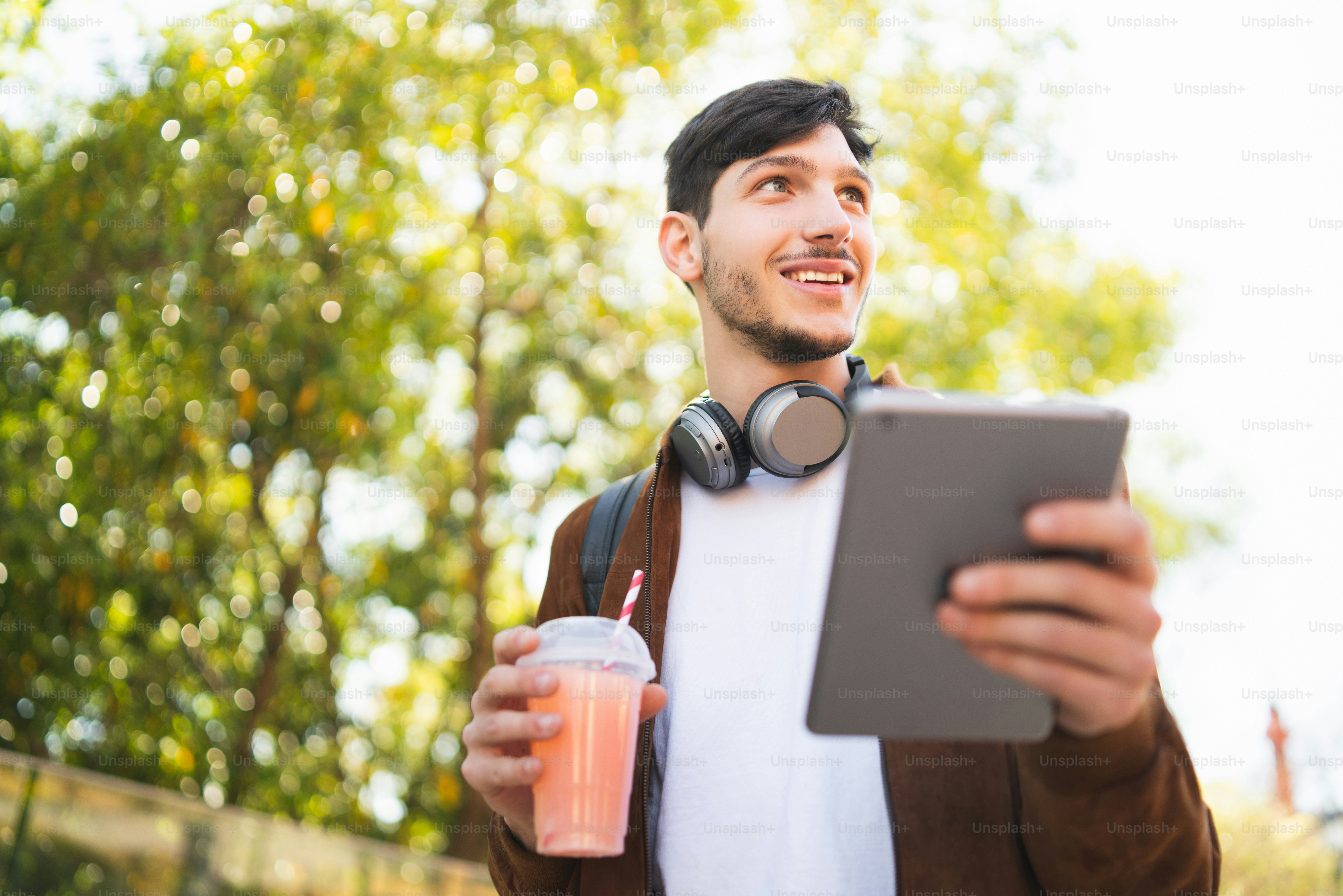 Portrait of young man using his digital tablet while drinking fresh fruit juice outdoors in the street. Technology and urban concept.