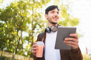 Portrait d’un jeune homme utilisant sa tablette numérique tout en buvant du jus de fruits frais à l’extérieur dans la rue. Technologie et concept urbain.