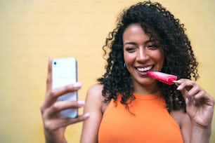 Ritratto di donna afro che scatta selfie con il suo telefono mophile mentre mangia un gelato. Tecnologia e concetto di lifestyle.