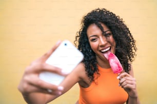 アイスクリームを食べながらモフィルの携帯電話で自分撮りをするアフロ女性のポートレート。テクノロジーとライフスタイルのコンセプト。