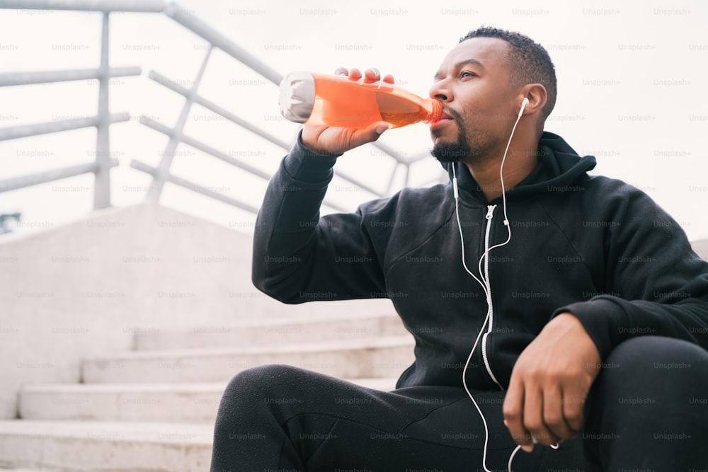 Retrato de un hombre atlético bebiendo algo después de entrenar mientras está sentado en escaleras de concreto. Deporte y estilo de vida saludable.