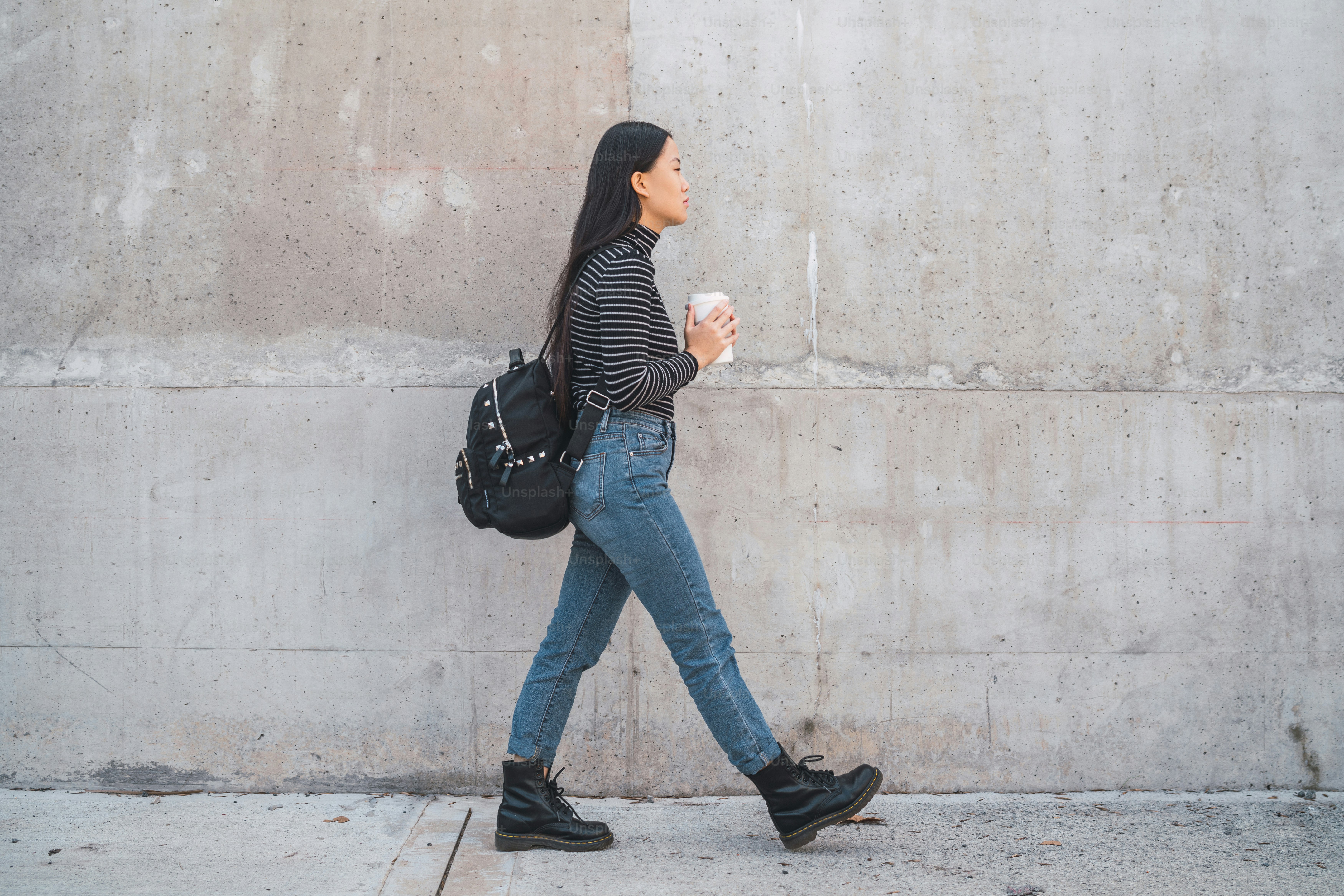 Portrait of young Asian woman walking and holding a cup of coffee against grey wall.