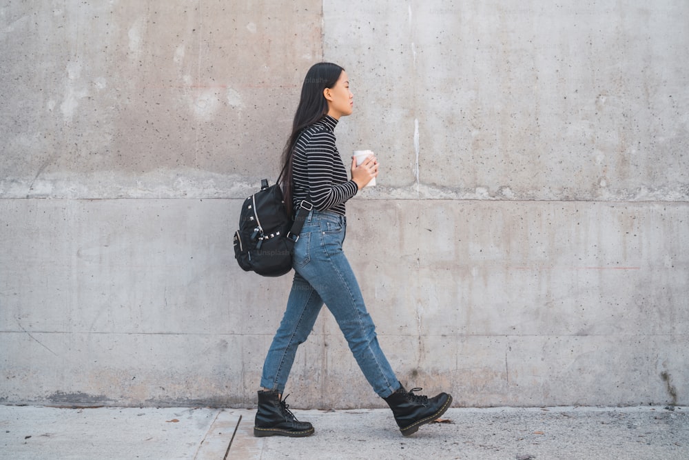 Retrato da jovem mulher asiática andando e segurando uma xícara de café contra a parede cinzenta.