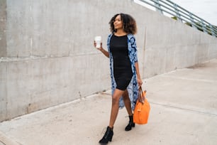 젊은 아프리카계 미국인 여성의 초상화는 야외에서 커피 한 잔과 가방을 들고 걷고 있다.