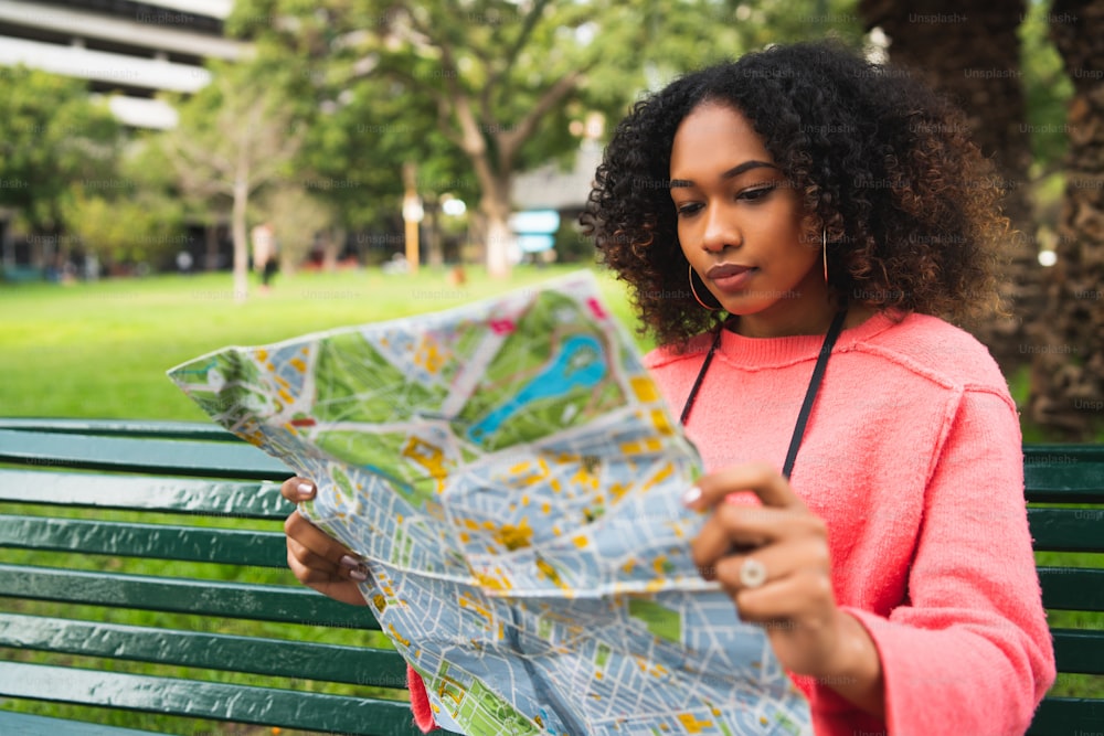 Retrato da jovem mulher afro-americana bonita sentada no banco no parque e olhando para um mapa. Conceito de viagem. Ao ar livre.