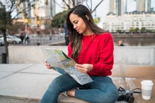 地図を持ち、通りで屋外の道順を探している若いラテン系の女性のポートレート。旅行のコンセプト。