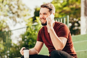 Young man sitting in public park drinking coffee and talking on cellphone