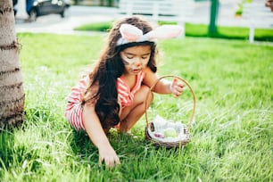 Cute little girl with bunny ears and basket of Easter eggs in the garden. Easter egg hunt