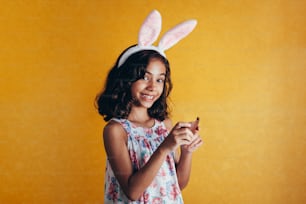 Criança pequena bonito usando orelhas de coelho no dia de Páscoa no fundo da cor. Menina que come o ovo de páscoa do chocolate