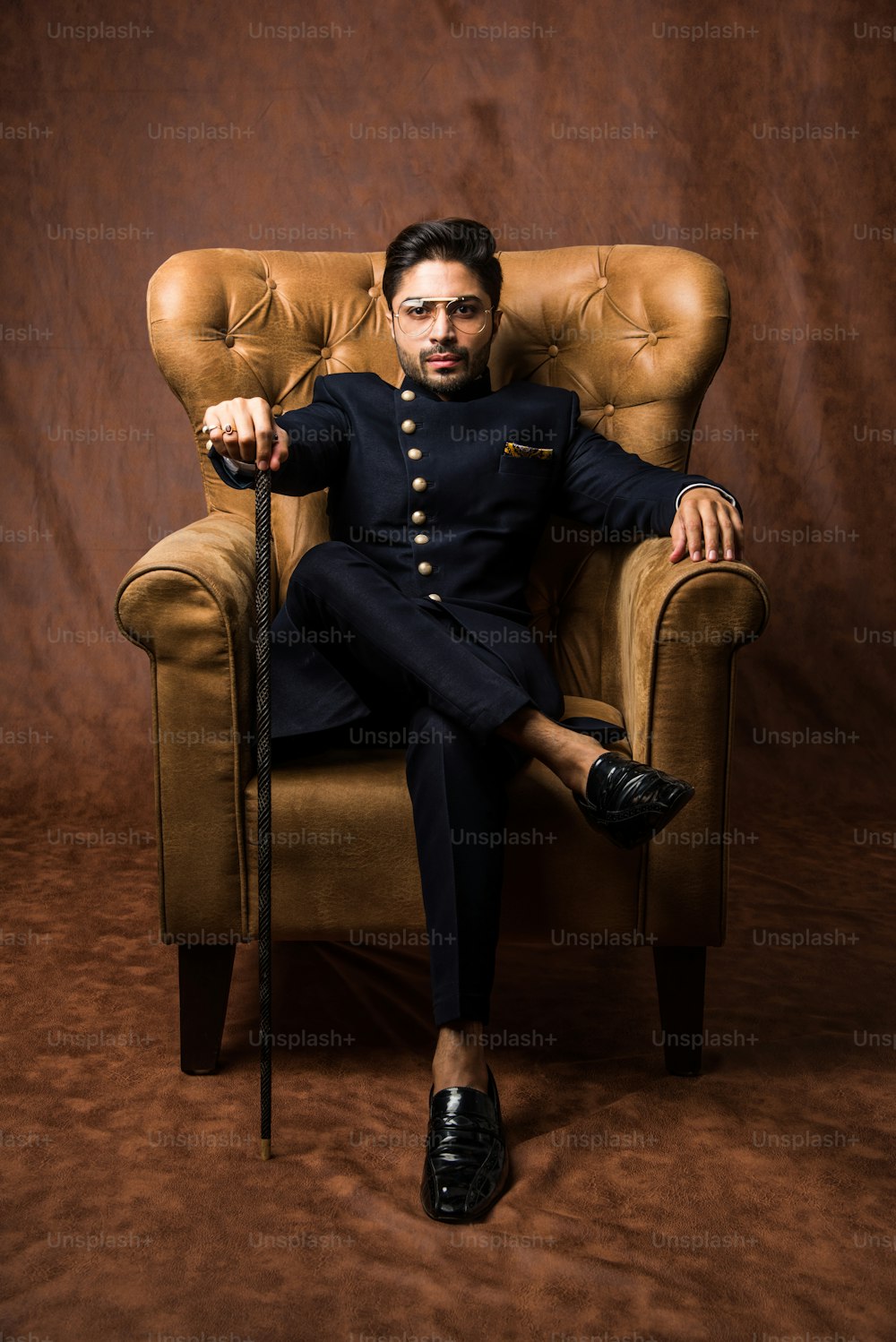 Indian bridegroom wears ethnic or traditional cloths,  Male fashion model with dark blue sherwani, Sitting on Wing Chair / sofa over brown grunge background, selective focus